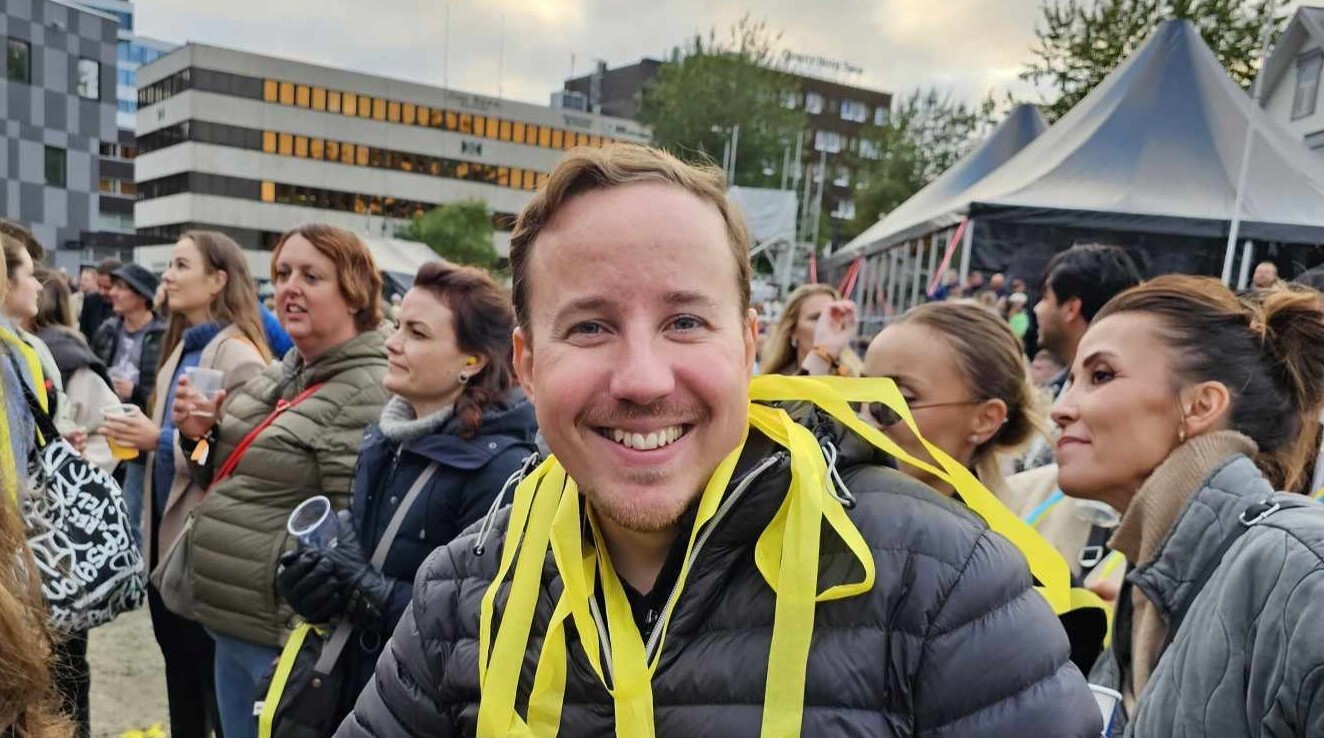 Mann i forgrunn med festivalgjester i bakgrunn, han har gule papirremser hengende over skuldrene