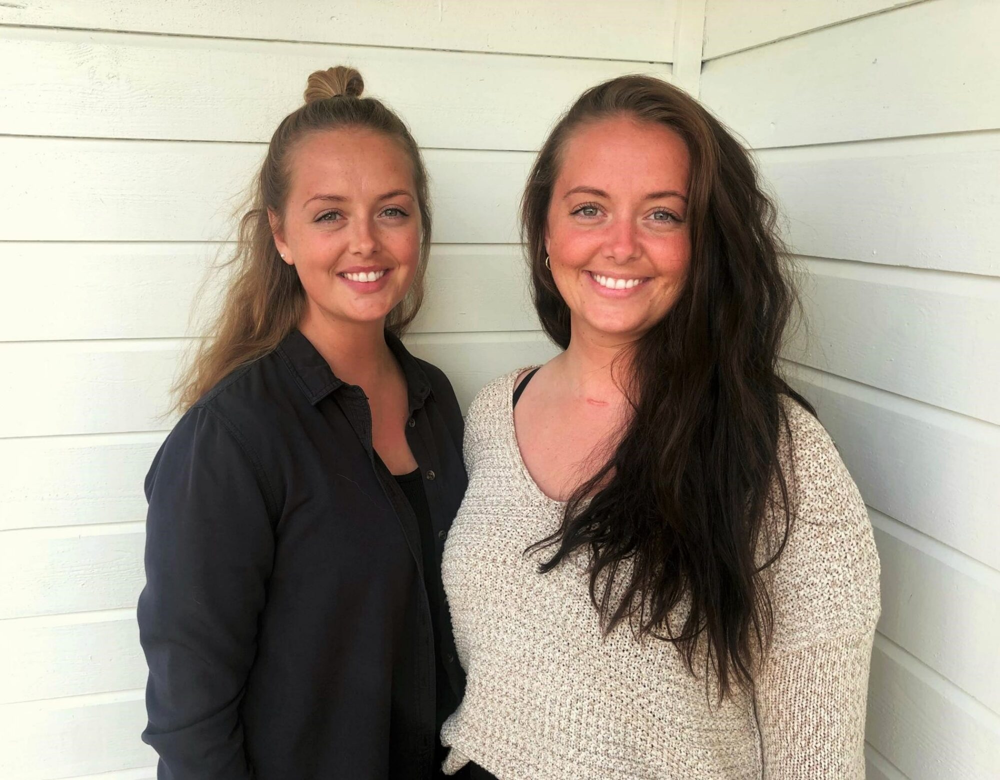 Camilla og Caroline står inntil en husvegg og smiler i kamera