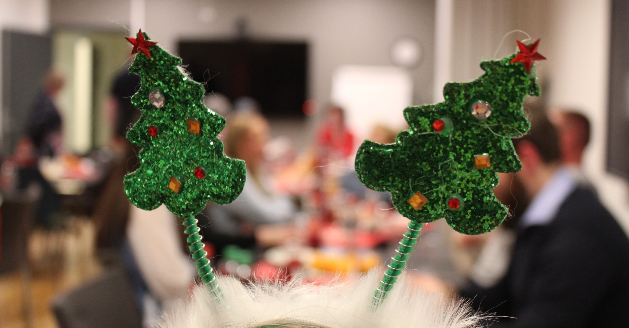 I fokus er en hårbøyle med to glitrende juletrær på. I bakgrunnen ser vi medlemmer som spiser grøt