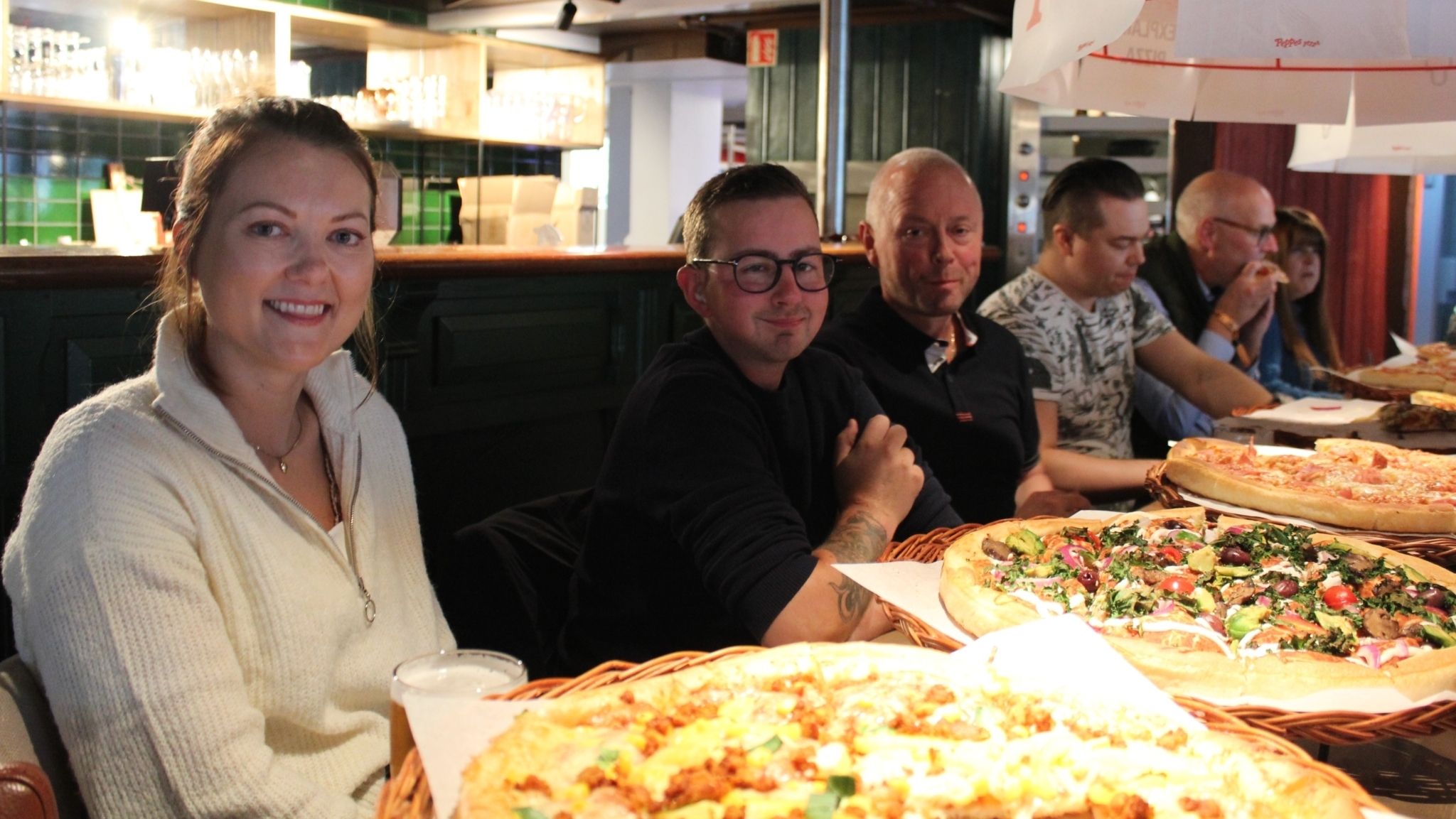 Fem personer sitter langs et bord, med pizza nedover bordet