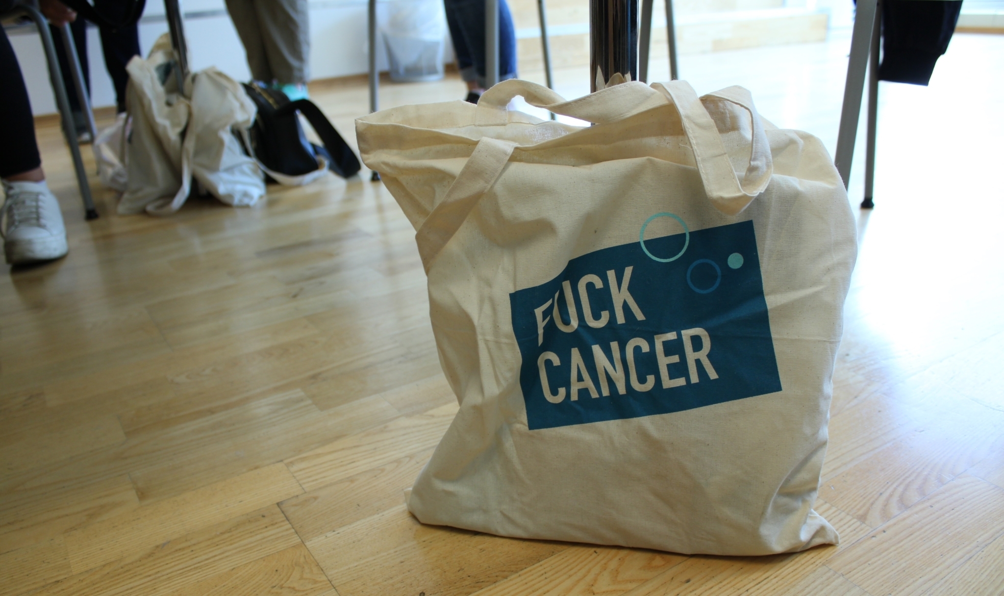 Et Fuck Cancer-bærenett med hvit tekst på blå firkant står på et gulv. I bakgrunn skimtes stolbein og føtter med sko på.