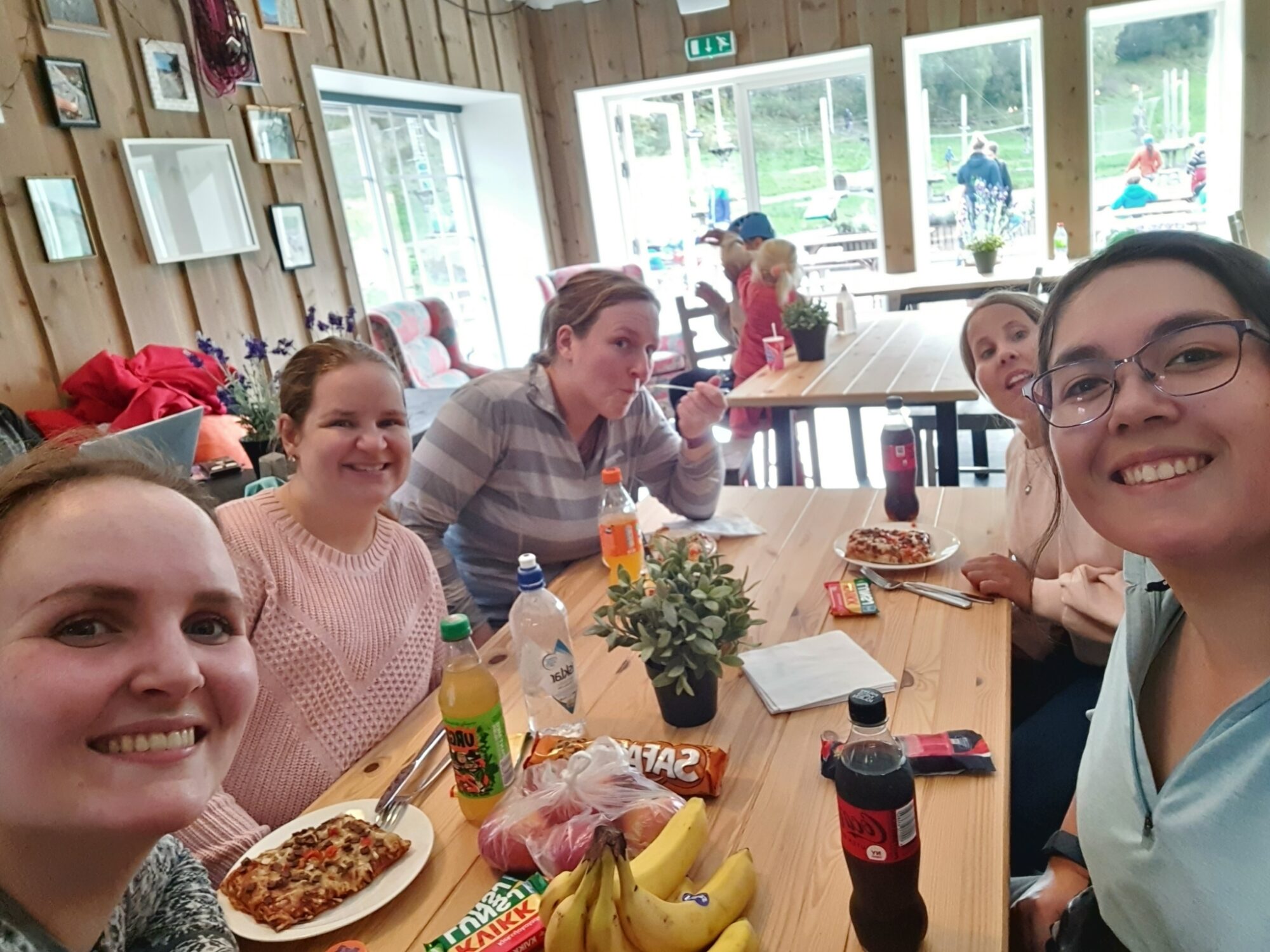 Fem unge kvinner sitter rundt et bord og spiser lunsj mens de smiler inn i kamera.