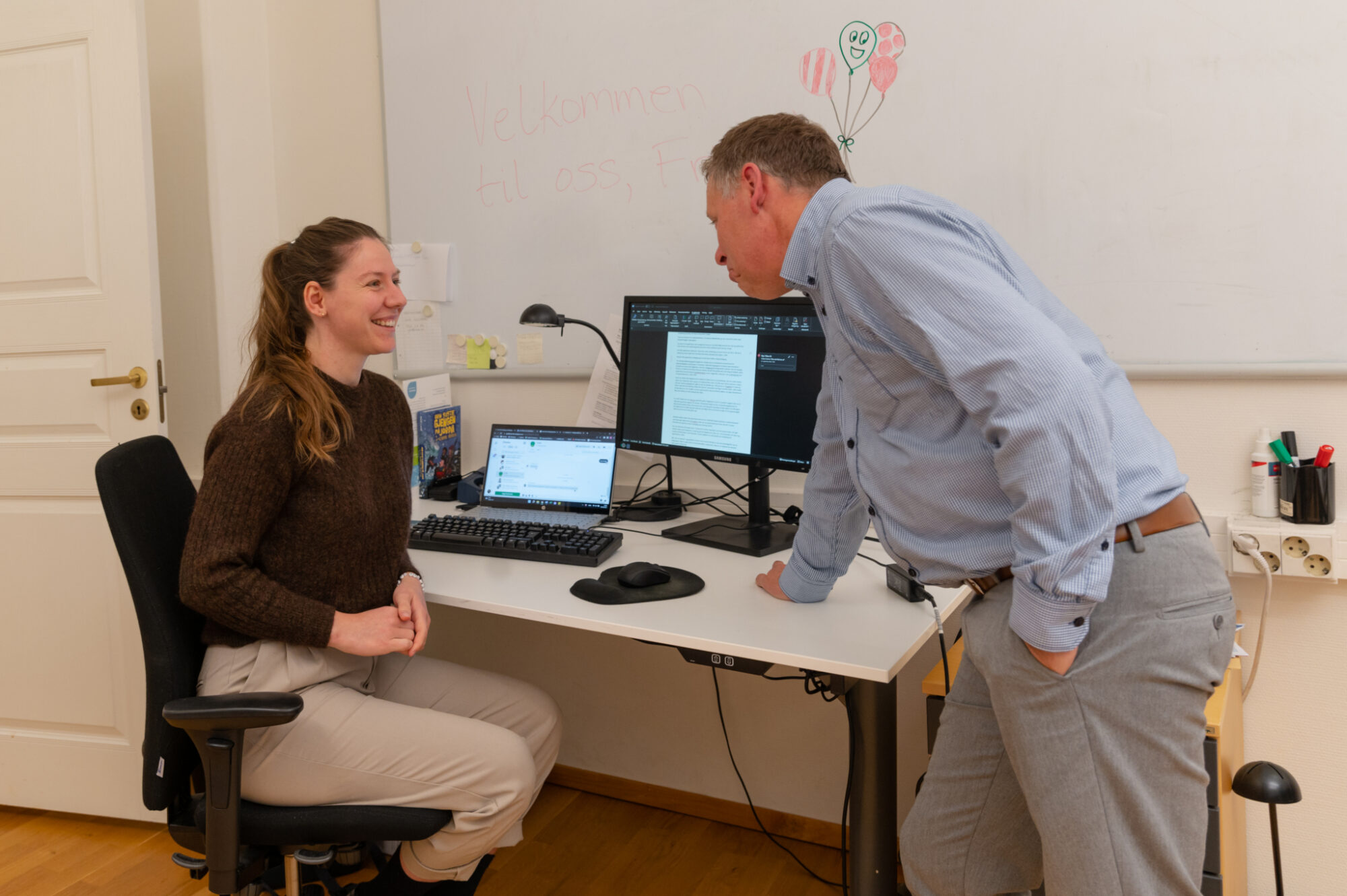 Frida sitter ved arbeidsplassen sin med pc og andre arbeidsverktøy foran seg. Sjefen hennes lener seg over pulten og snakker med henne. De smiler til hverandre.